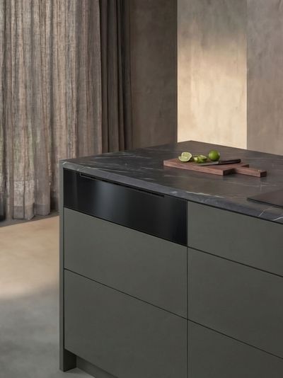&Icirc;lot de cuisine gris mat &agrave; plan marbre et panneau noir, planche avec citrons verts, rideau laissant entrevoir un jardin et table dress&eacute;e en arri&egrave;re-plan.