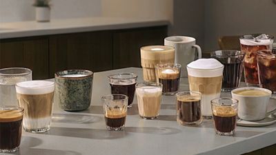 Hombre en su&eacute;ter verde de pie en la cocina, sosteniendo taza de caf&eacute;, m&aacute;quina de caf&eacute; en el mostrador.