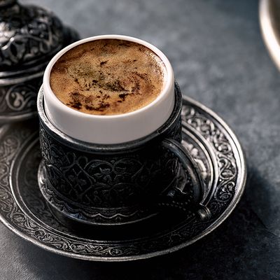 Frischer türkischer Kaffee in einer orientalisch gestalteten Tasse mit Deckel und türkischem Gebäck im Hintergrund