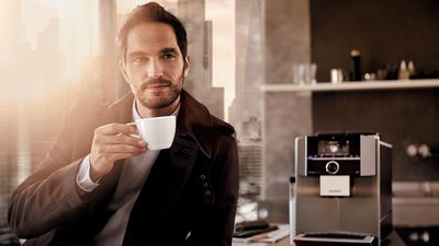 Ein Mann trinkt einen Kaffee in der Küche. Auf dem Tisch steht ein Siemens EQ.9 Kaffeevollautomat