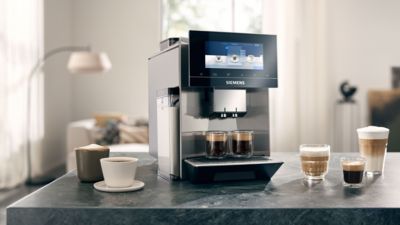 Une machine à café automatique Siemens EQ900 sur un comptoir de cuisine, entourée de spécialités de café, dont un cappuccino, un latte et un expresso.