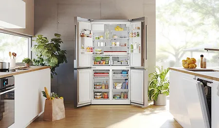 A modern kitchen featuring an open refrigerator stocked with fresh food and drinks, surrounded by greenery and bright natural light.