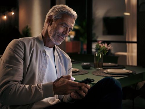 Ein älterer Herr sitzt am Abend am Esstisch und liest etwas auf seinem Smartphone.