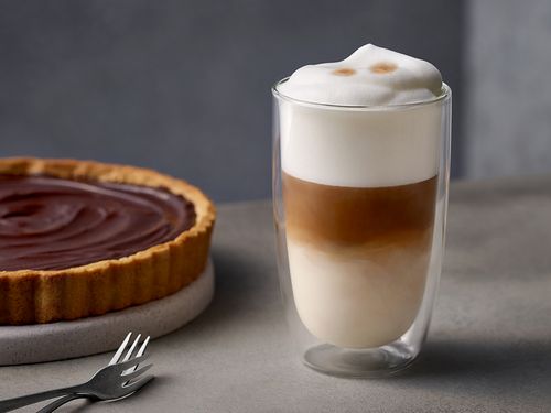 Ein isoliertes Glas mit Latte Macchiato, mit einem Kuchen und einem Zuckerschälchen im Hintergrund
