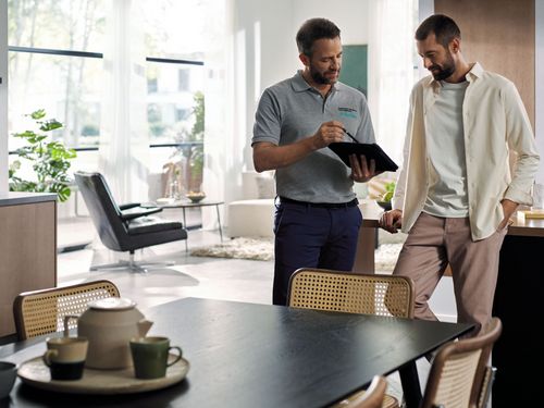 Twee mannen discussi&euml;ren over een tablet in een moderne keuken met planten, stijlvolle meubels en een gedekte eettafel.