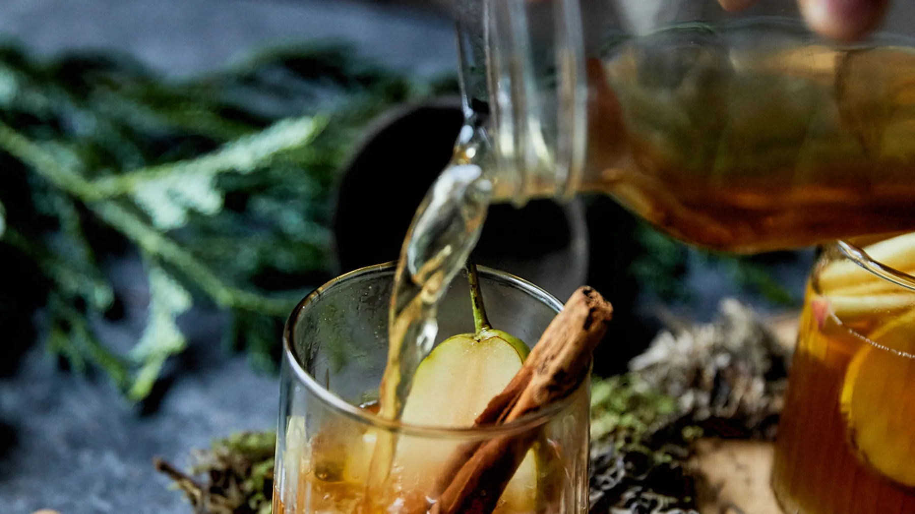 Bicchiere con bevanda chiara, un bastoncino di cannella e una fetta di mela, con liquido versato da una brocca