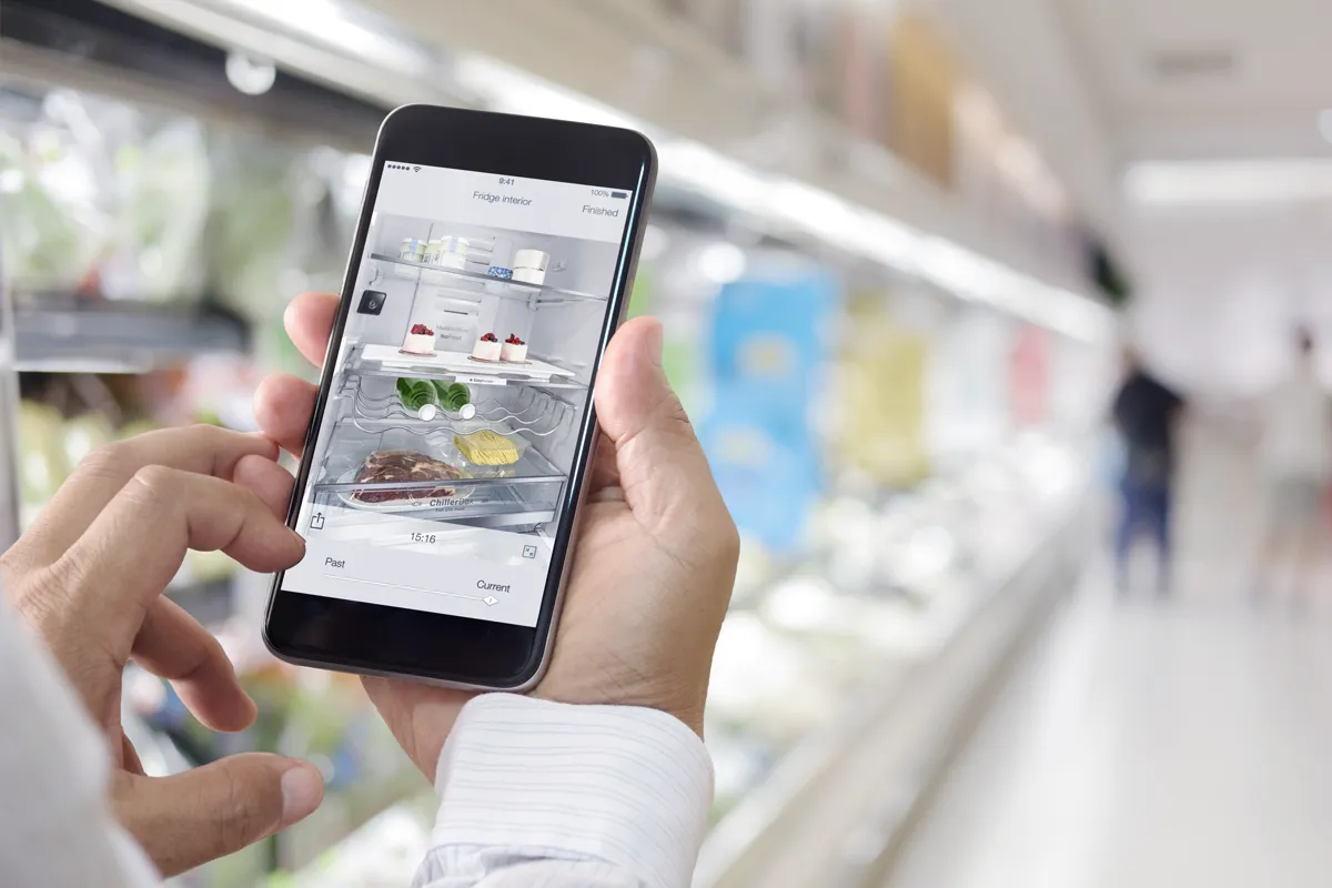 Hands holding smartphone in a supermarket. On the screen is smart fridge's interior.