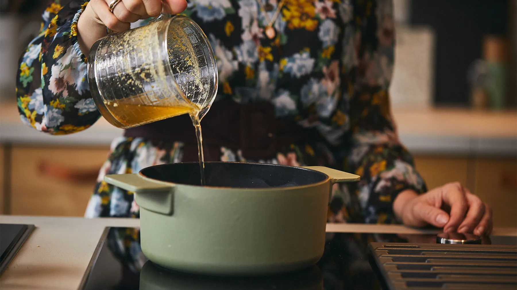 Donna versando liquido da vaso di vetro in pentola su stufa, utensili da cucina visibili.