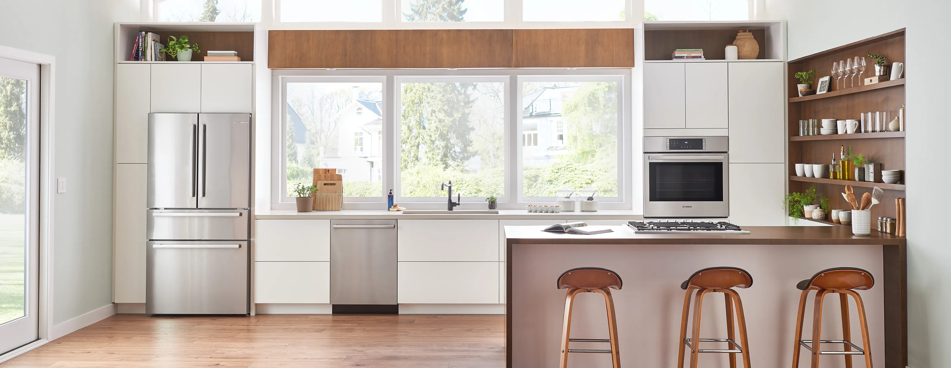 Modern kitchen with Bosch stainless steel appliances, large windows, and wooden bar stools.