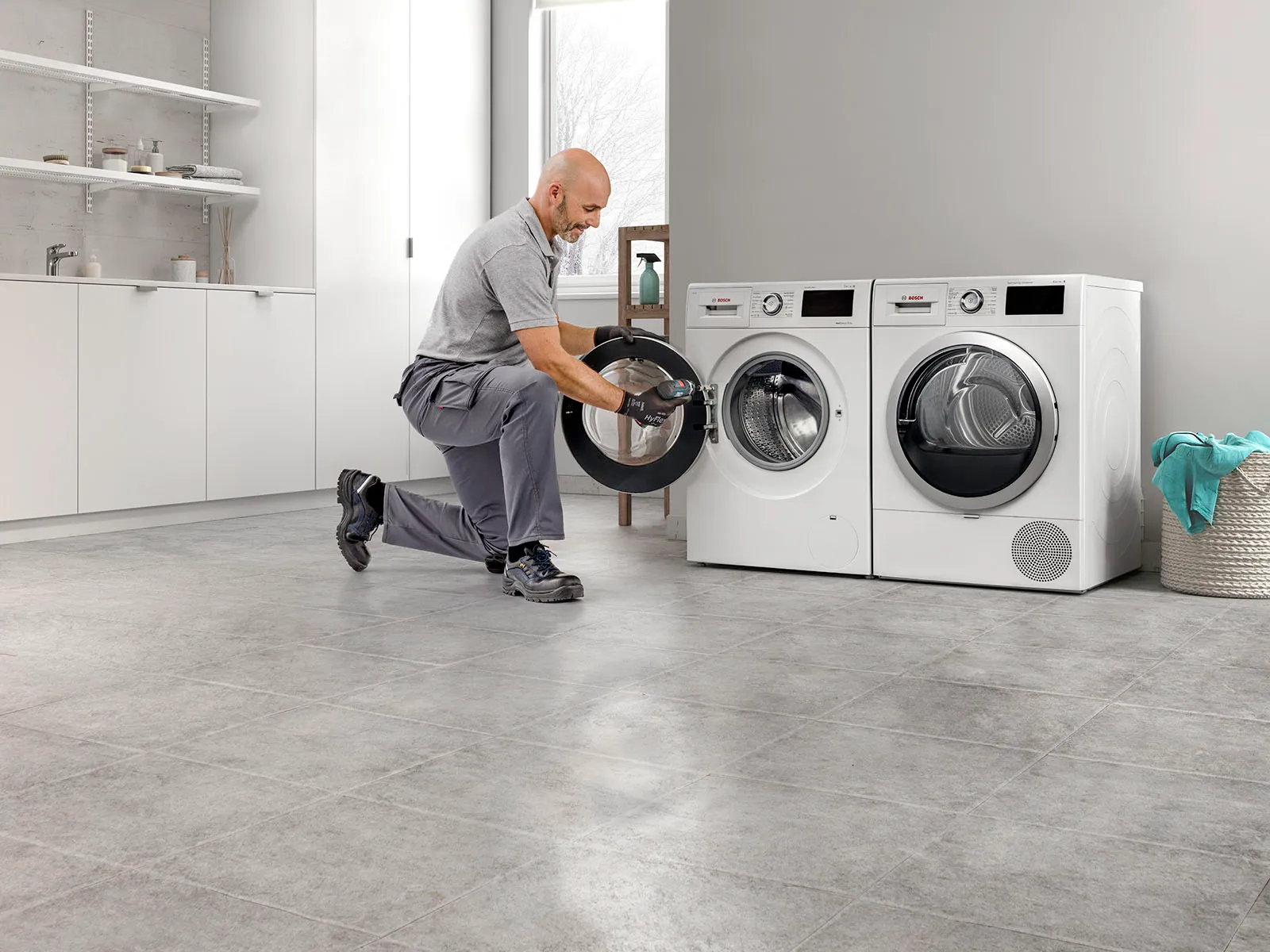 Man fixing washing machine in modern laundry room with shelves and appliances.
