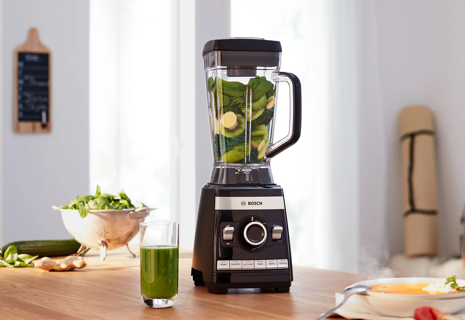 Blenderul Bosch Vitaboost de mare viteză, cu spanac și banană în interior, așezat pe blatul de bucătărie lângă un pahar cu smoothie verde și un bol cu salată