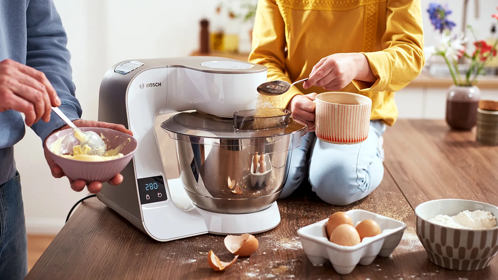 Scena de bucatarie cu persoana folosind un mixer stand pentru a pregăti produse coapte, înconjurat de ingrediente.