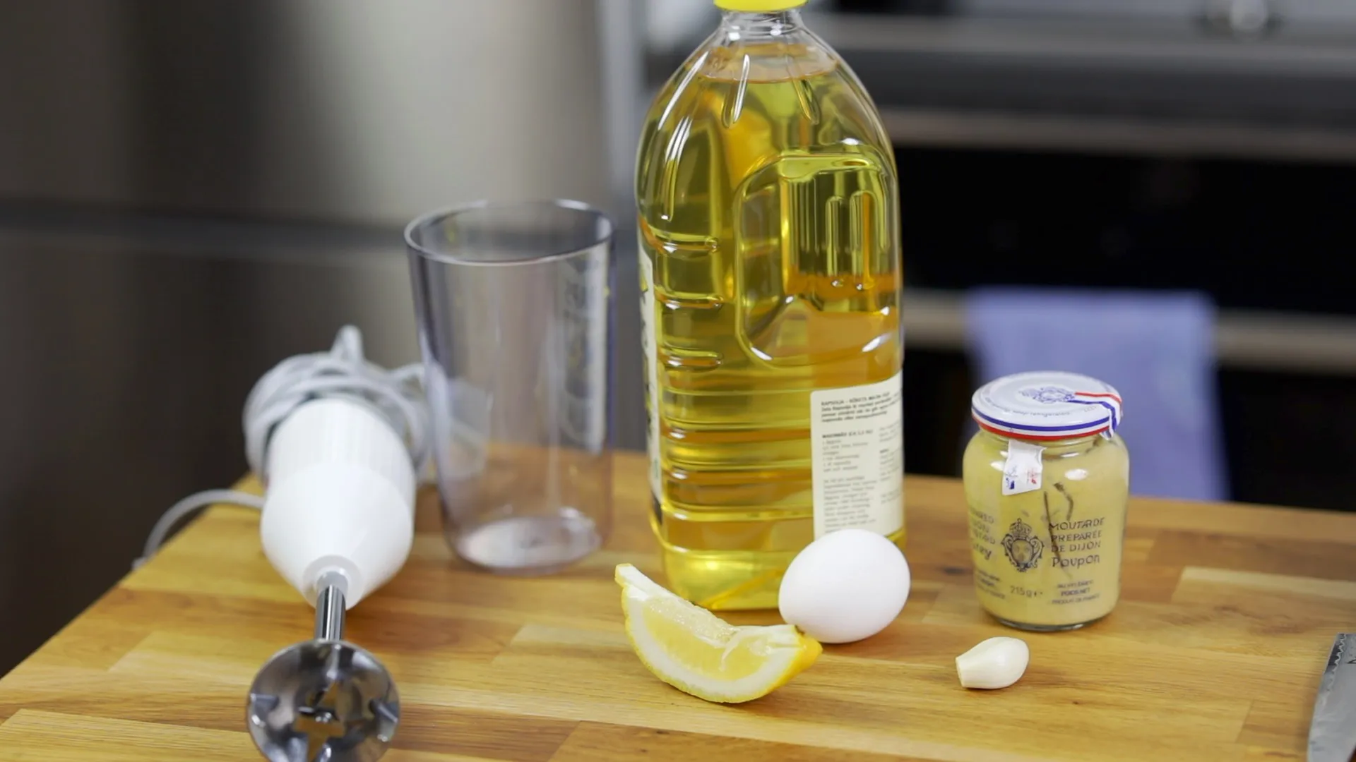 En köksarbetsyta med en stavmixer, ingredienser som ägg, citronsaft, olja och senap för en sås.