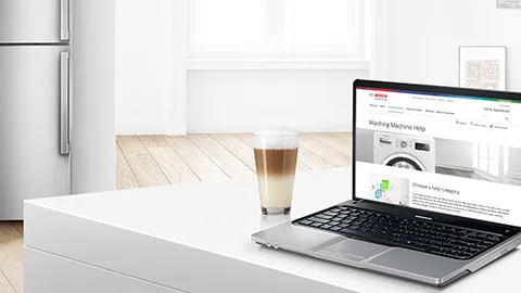 A laptop displays a Bosch washing machine help page beside a cup of coffee on a modern kitchen table. Bright, airy interior design.
