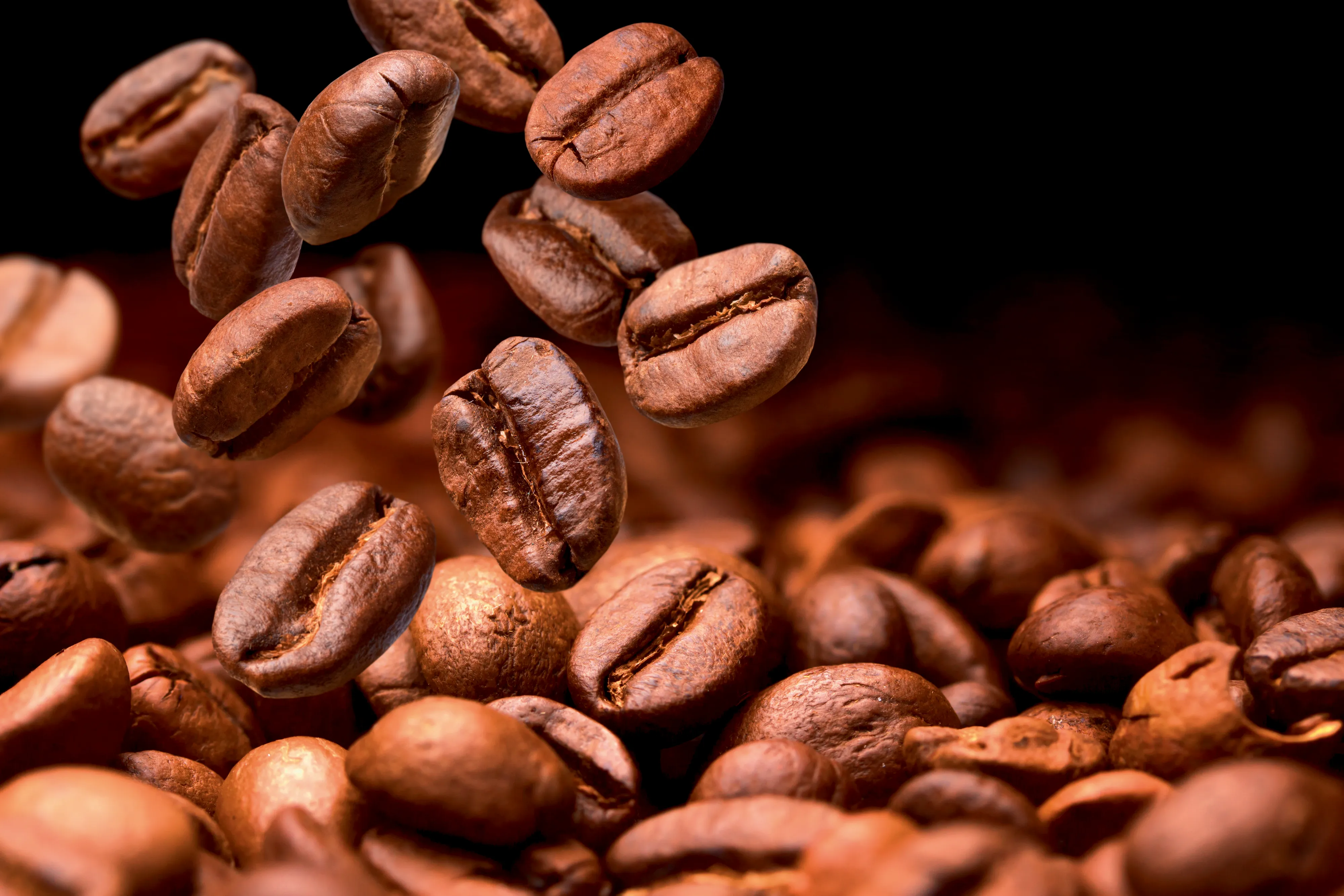 Close-up of roasted coffee beans in various shades of brown, some floating above a bed of beans against a dark background.