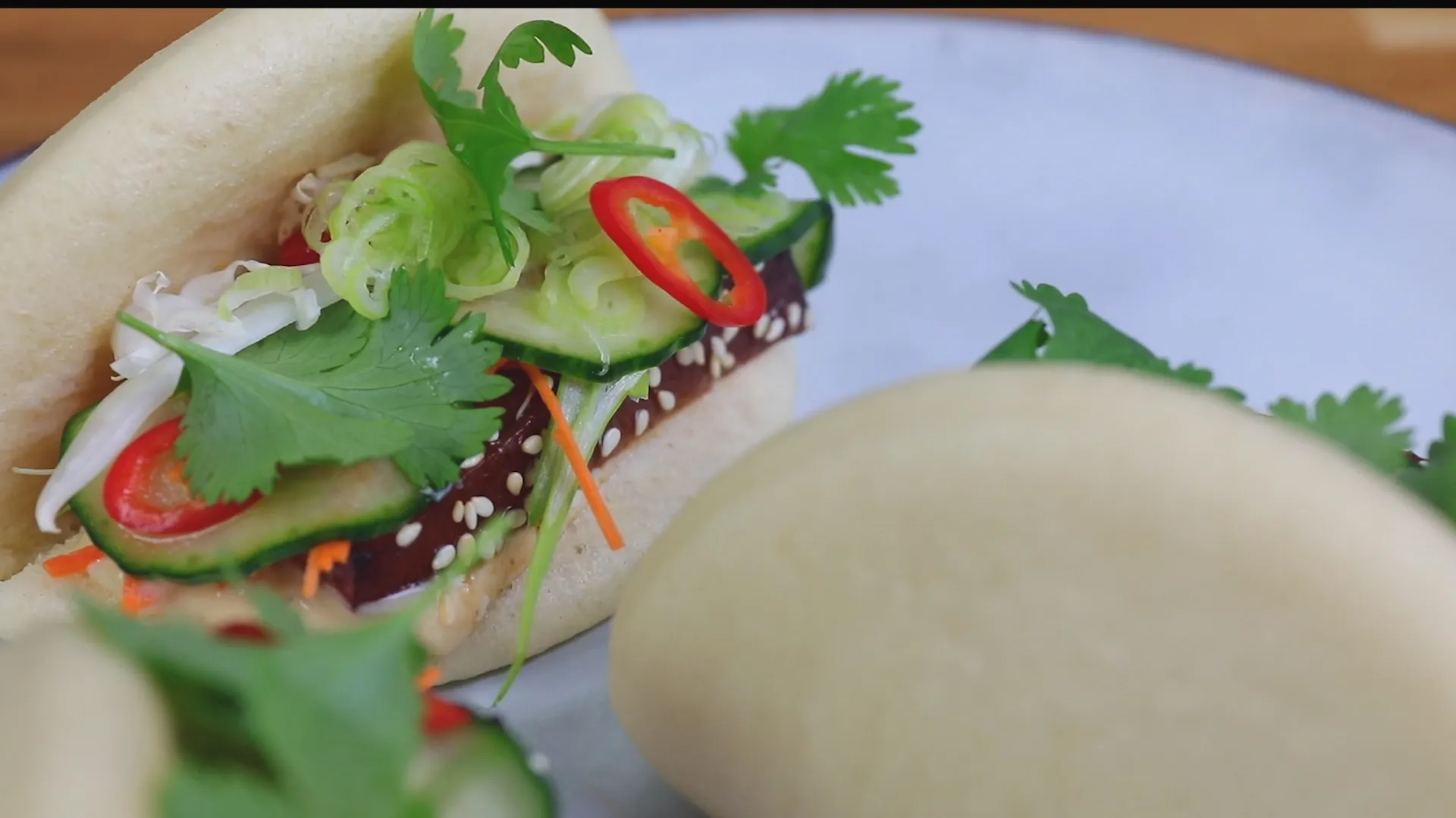 En fylld bao-bulle med krispiga grönsaker, koriander och skivor av chili på en tallrik, omgiven av andra bulle.