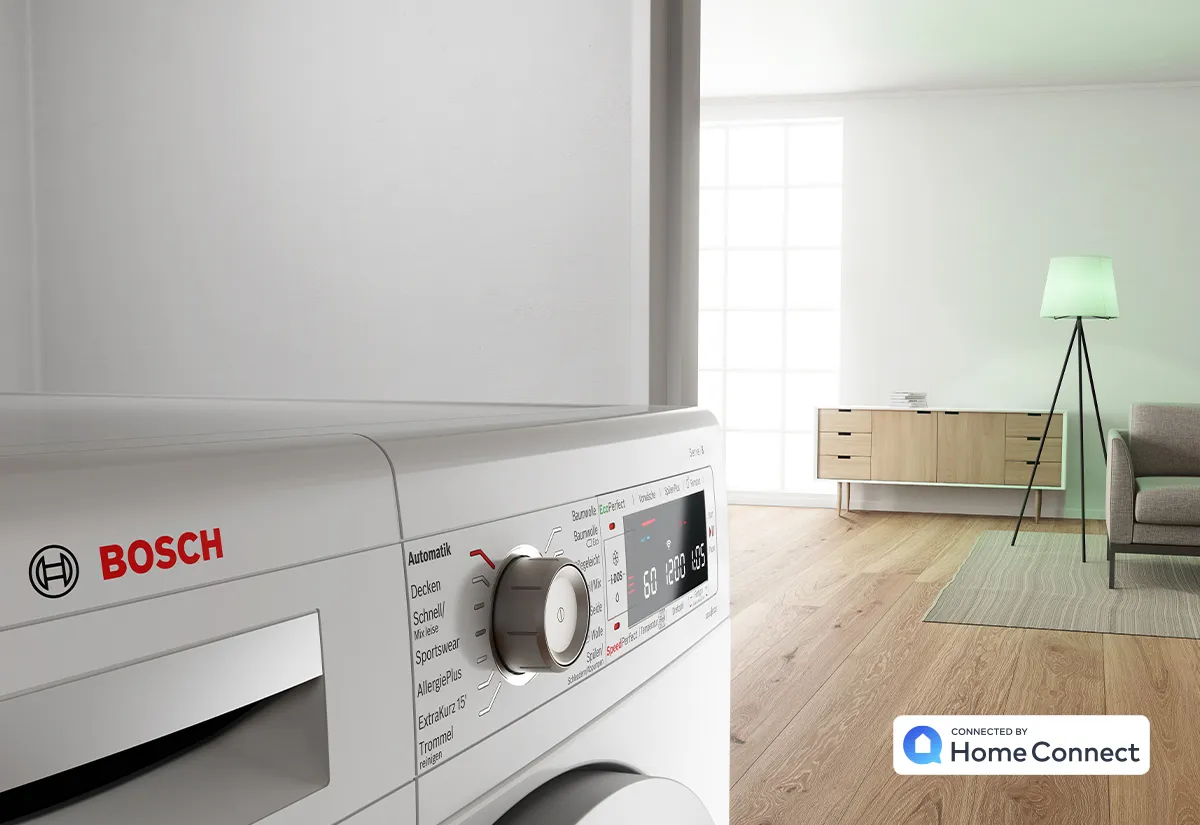 Close-up of a Bosch washing machine in a bright, modern living room with wooden floor, sideboard, lamp, and sofa visible.