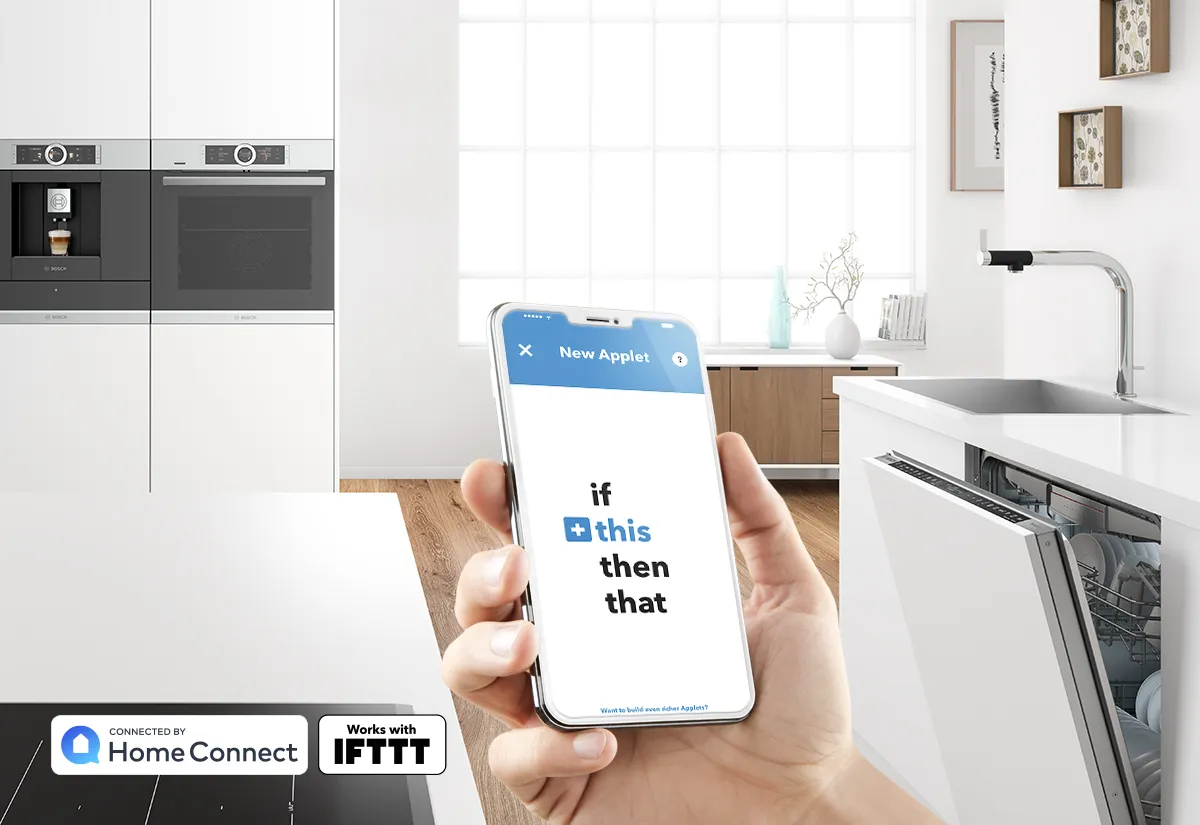 Hand holding a smartphone with an IFTTT applet screen in a modern white kitchen featuring a dishwasher and built-in appliances.