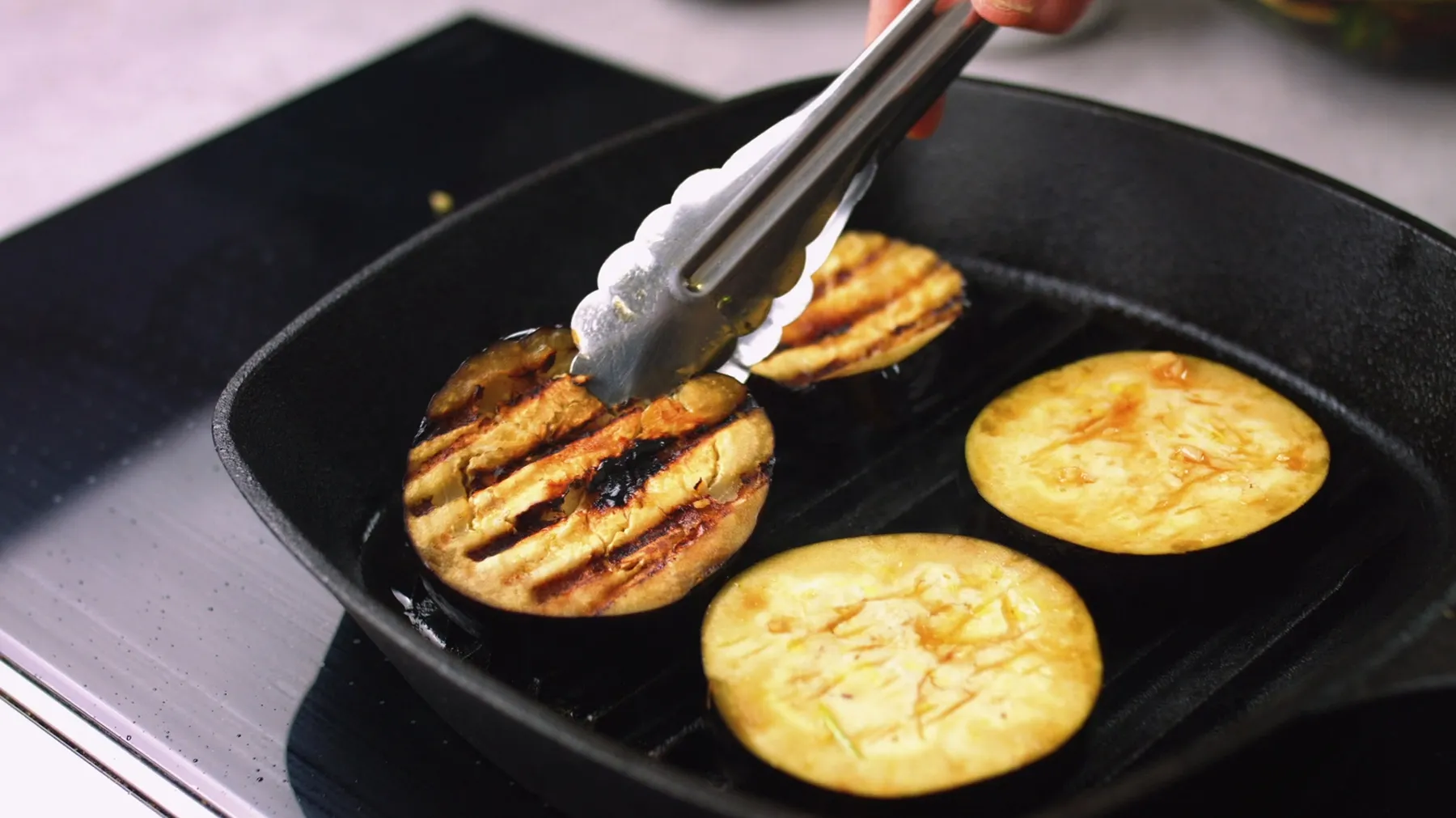 A hand uses tongs to flip a round slice of eggplant on a hot cast iron grill pan, showing prominent grill marks on the cooked side.