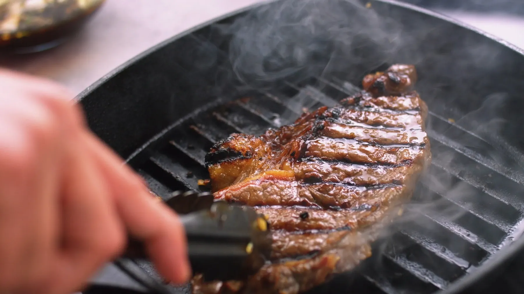 A hand uses tongs to flip a sizzling steak on a hot cast iron grill pan, creating visible smoke and grill marks.