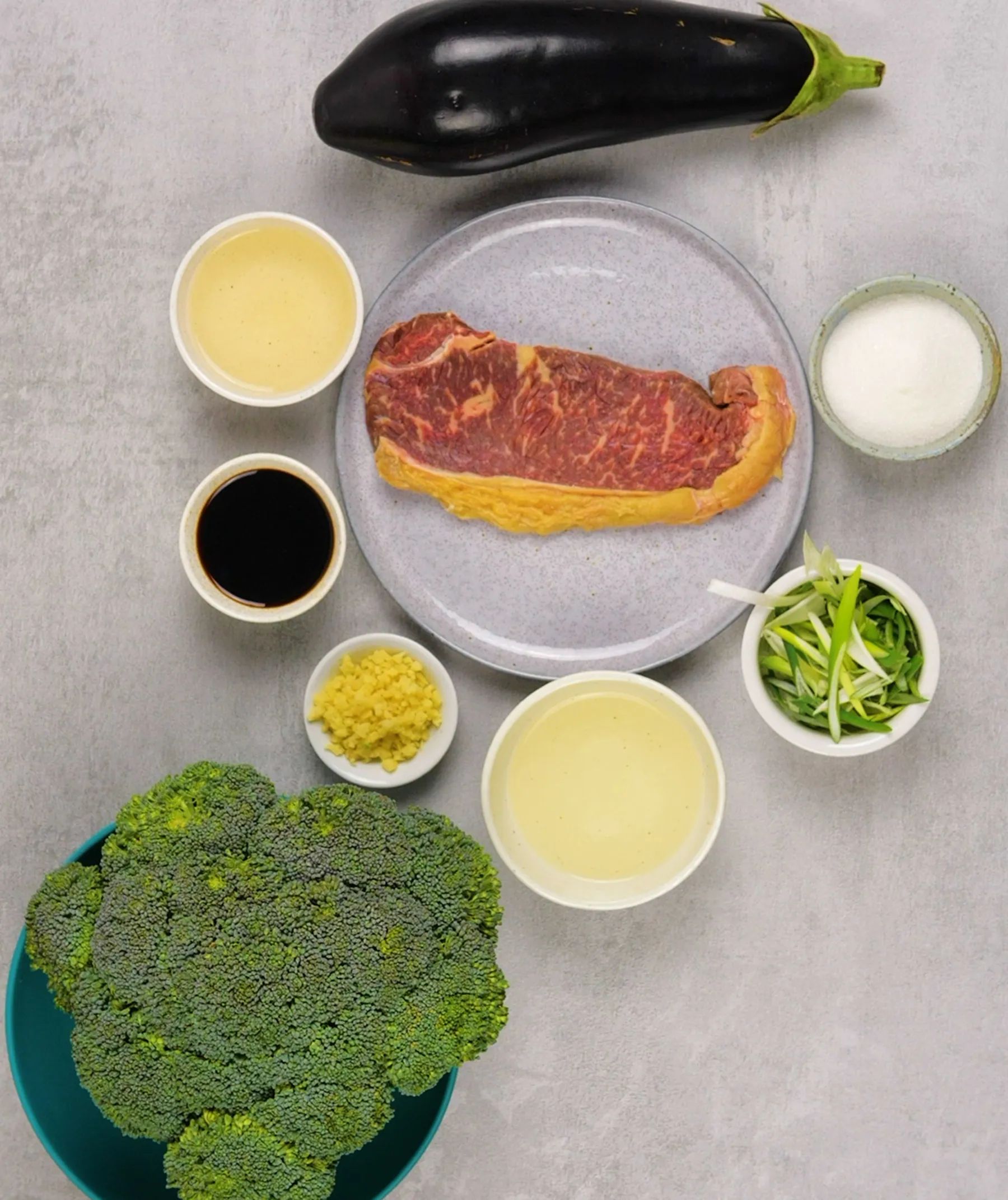 An overhead flat lay of ingredients for a meal: a raw steak, broccoli florets, an eggplant, bowls of oil, soy sauce, minced ginger, sugar, and sliced green onions, all on a grey background.