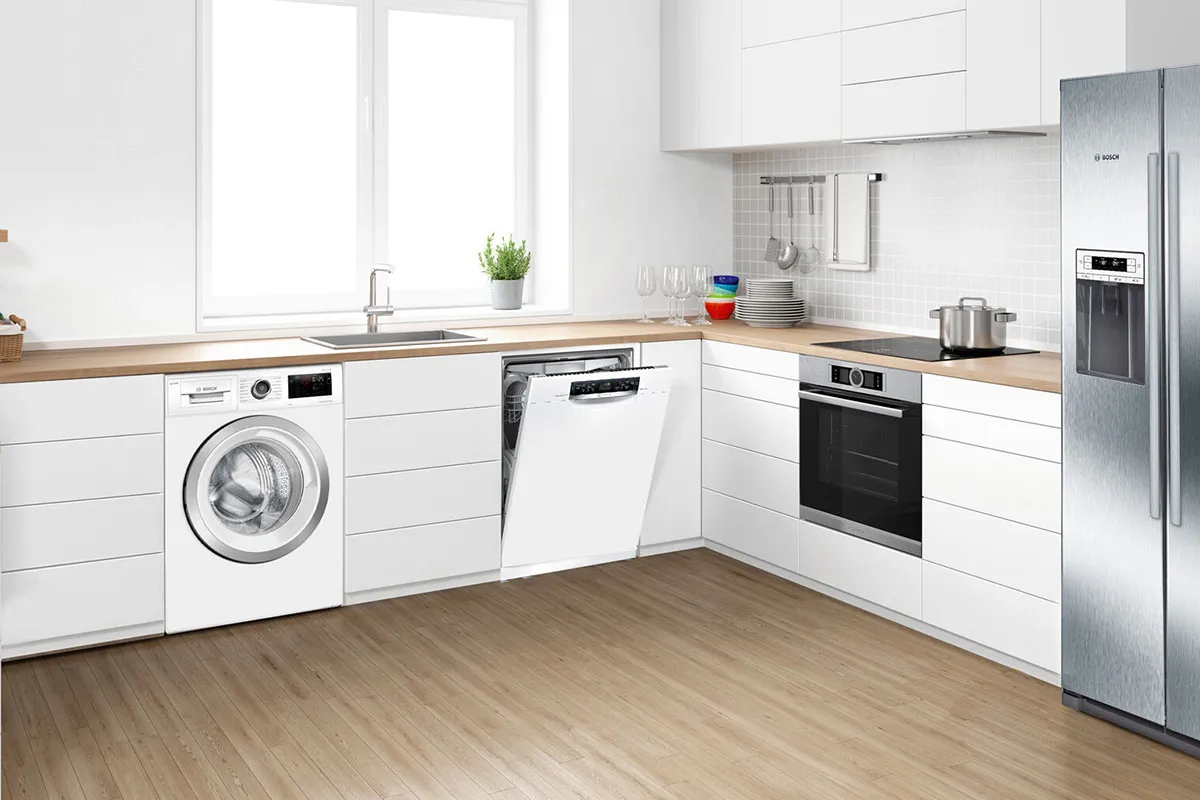 A wide shot of a modern kitchen featuring a row of Bosch appliances: a white front-loading washing machine, a white integrated dishwasher with the door ajar, a black built-in oven, and a large stainless steel American-style fridge-freezer.