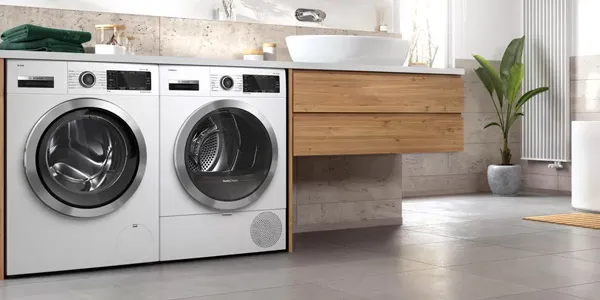 Lavanderia moderna con lavatrice e asciugatrice a carica frontale, mobile in legno, lavabo bianco e pianta in uno spazio luminoso e piastrellato.