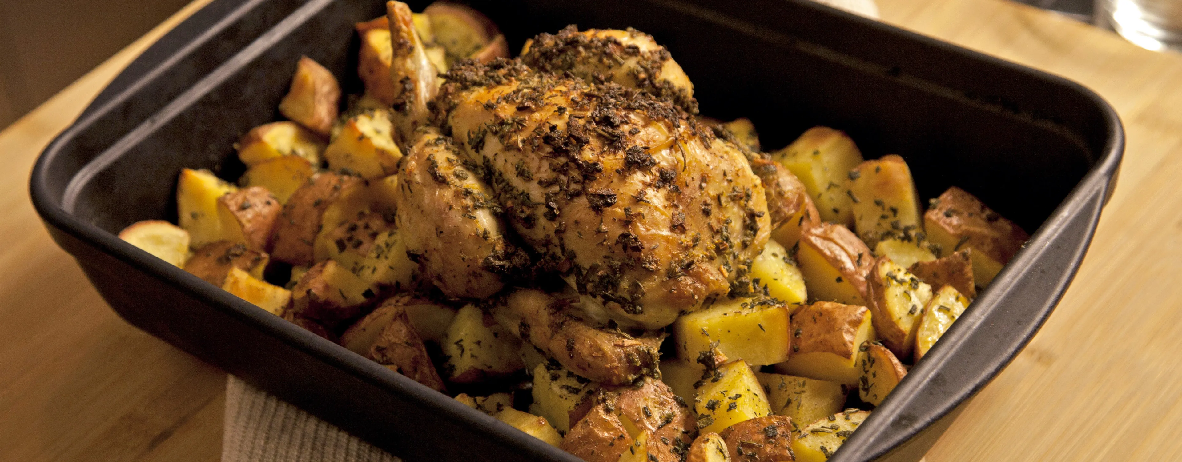 Pollo intero arrosto condito alle erbe, adagiato su pezzi di patate arrosto in una teglia nera appoggiata su un piano di legno.