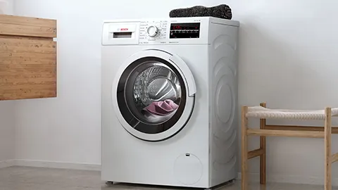 A modern white washing machine with a front-loading door, partially open, displaying pink laundry inside, next to a wooden chair.