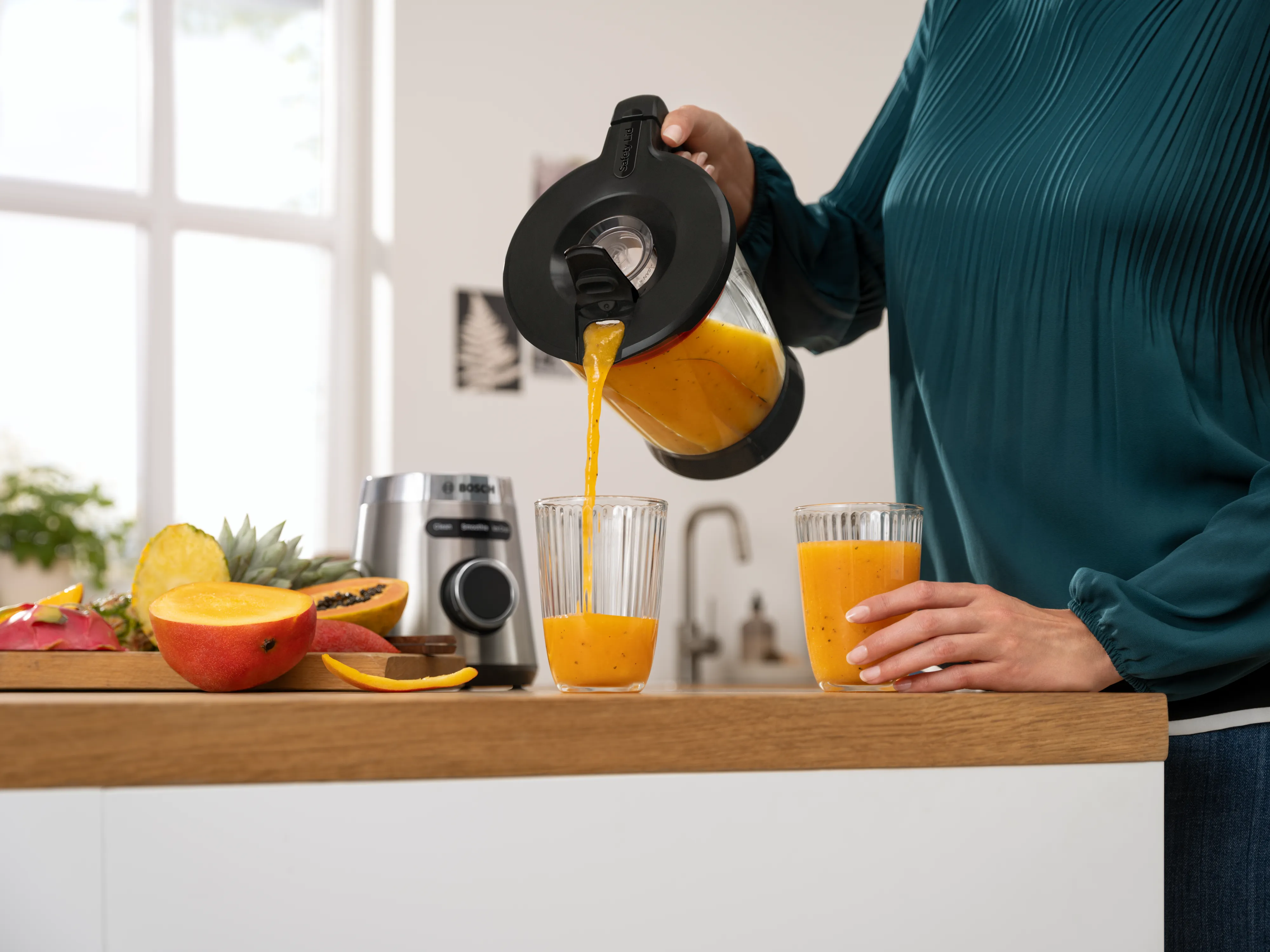 O persoană care toarnă suc de portocale dintr-un recipient de blender într-un pahar, pe blatul de bucătărie, cu fructe și baza blenderului în fundal
