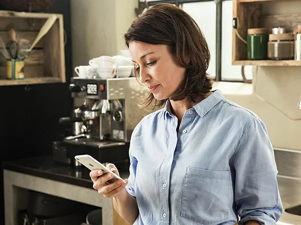 Donna in cucina con smartphone, abbigliamento casual, macchina da caffè in background.