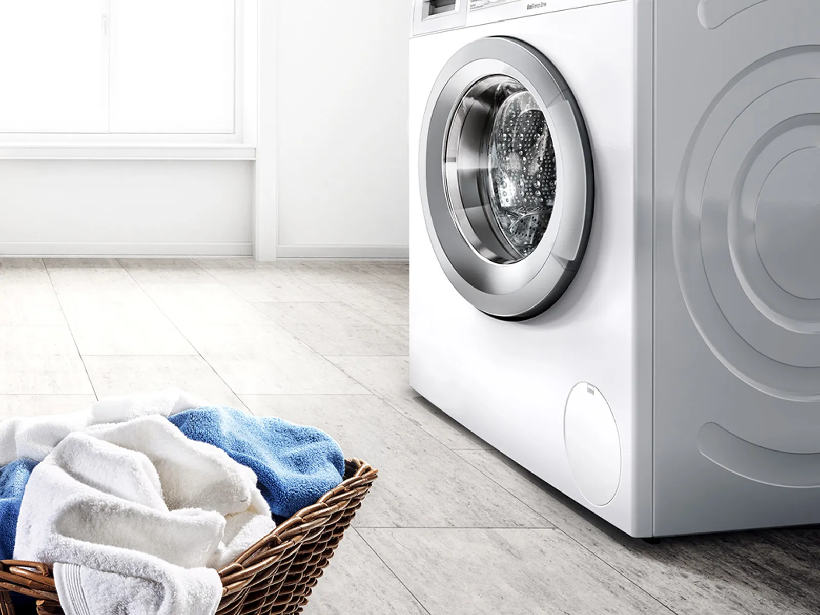 White front-loading washing machine on a light-tile floor by a sunlit window, with a wicker basket of folded towels beside it.