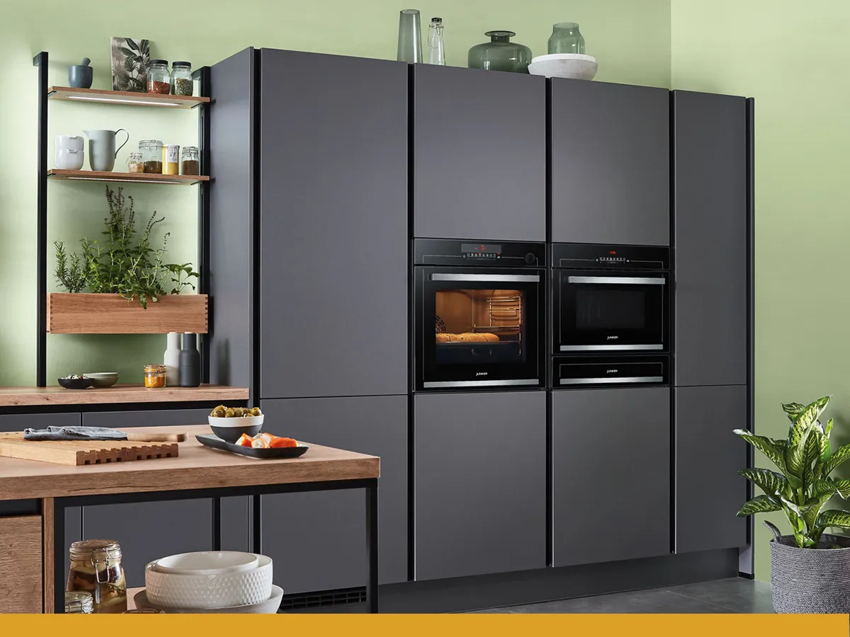 Moderne keuken met strakke grijze kasten, dubbele ovens, open planken met planten en een houten eettafel met servies.