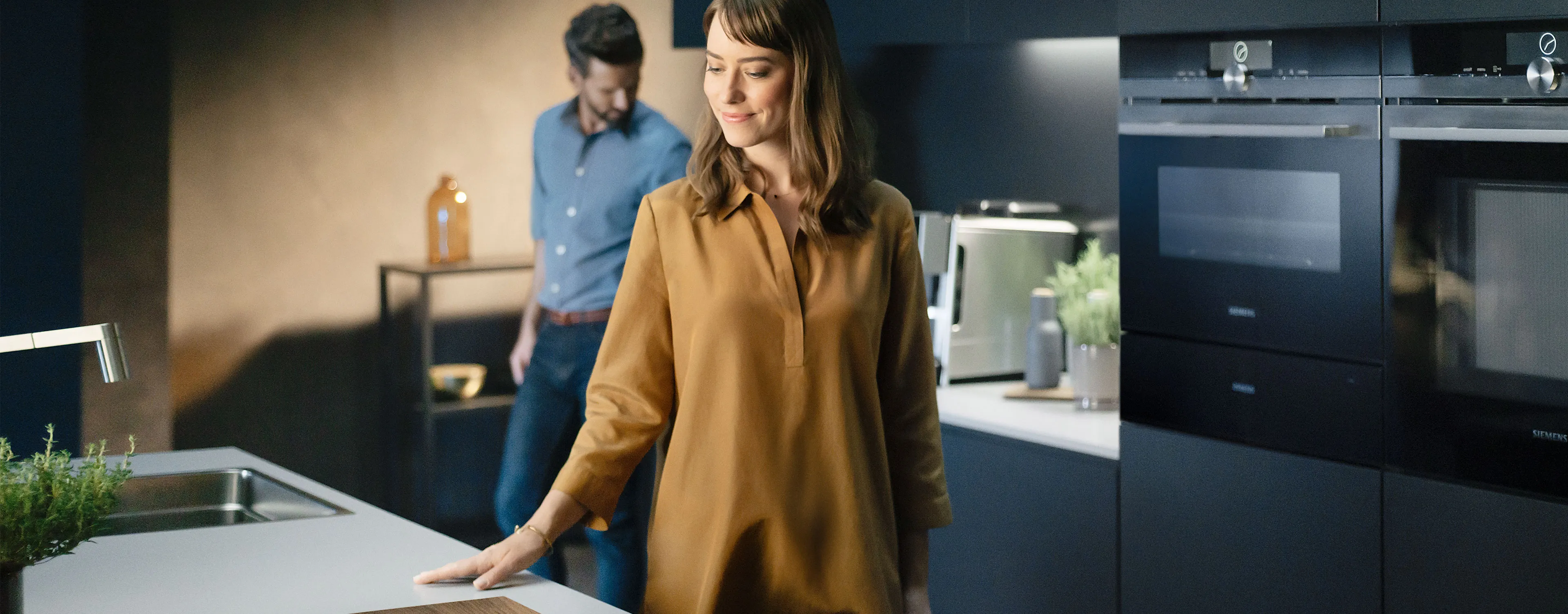 Woman in kitchen, man in background, modern appliances, warm lighting, cozy atmosphere.