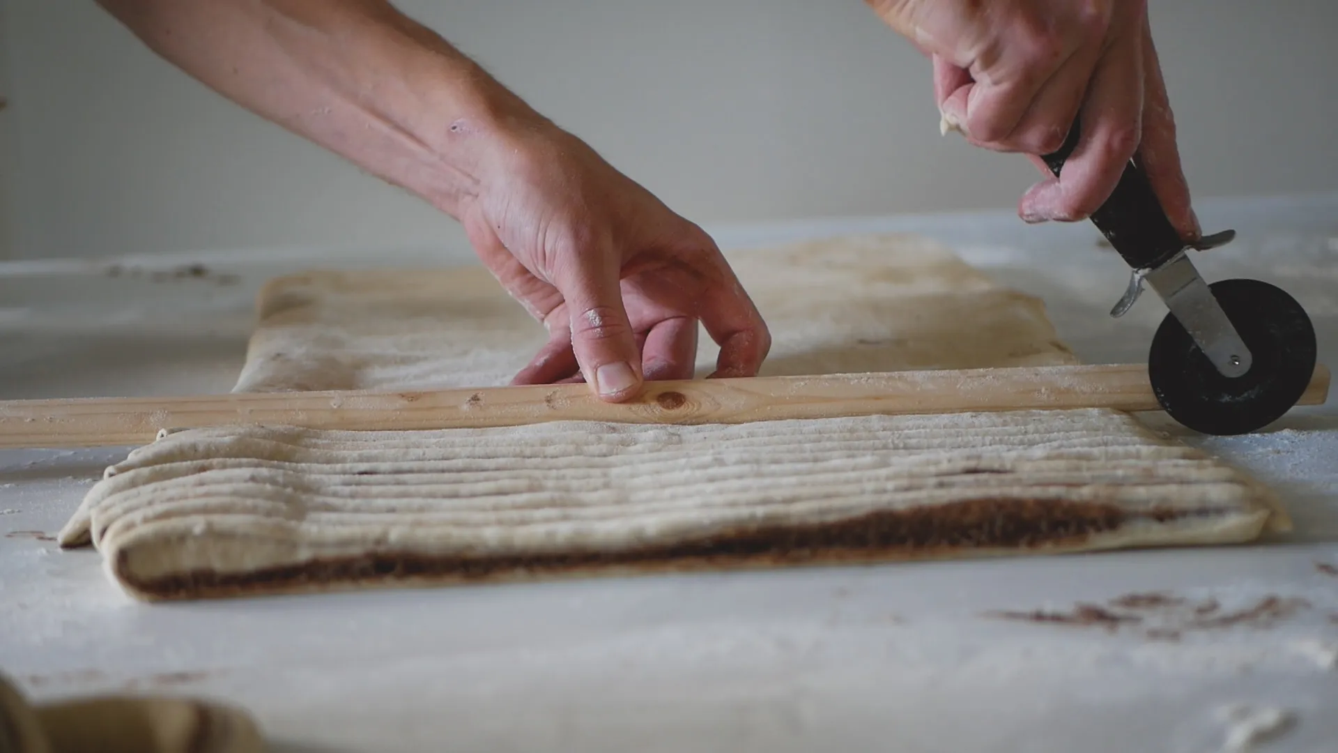 En matlagare använder en kavel och en pizzaskärare för att skära en deg med kanelfyllning på ett bänk.