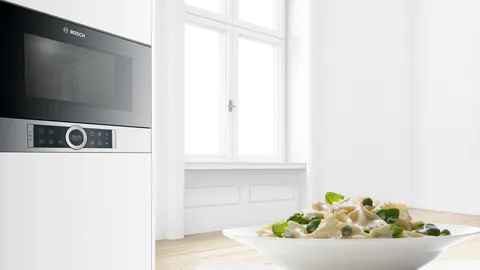 Modern kitchen with microwave, window, and bowl of fresh vegetables on wooden table.