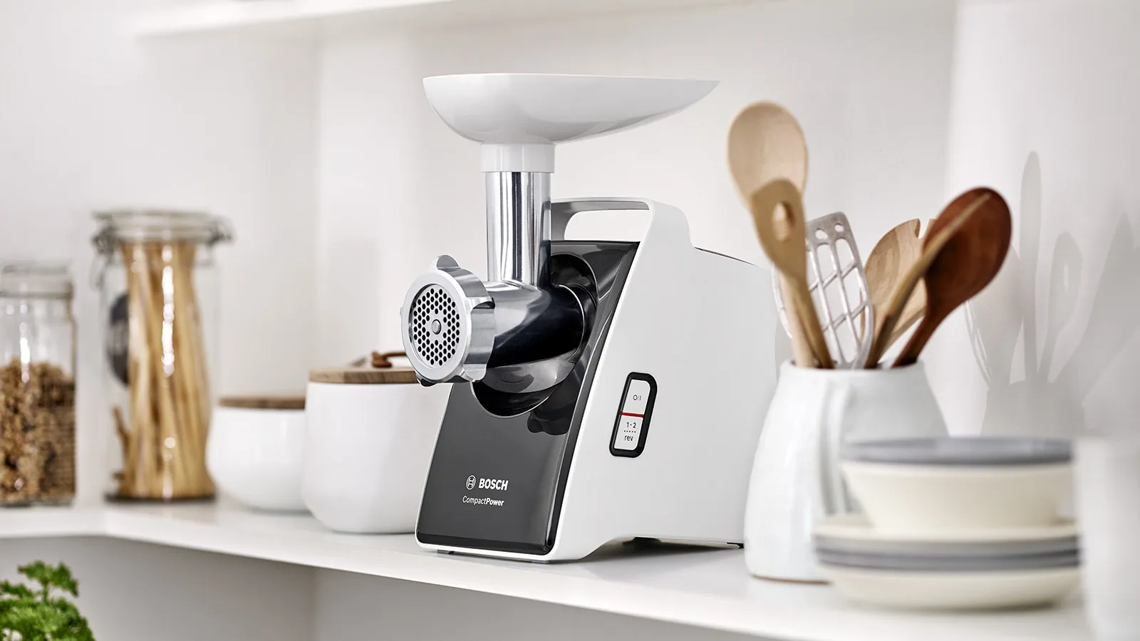 توجد مفرمة اللحوم Bosch CompactPower باللونين الأبيض والأسود على رف أبيض في المطبخ، وتحيط بها أدوات المطبخ والأواني.