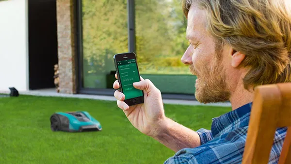 Hombre usando smartphone para controlar la cortadora de césped robótica en el patio trasero.