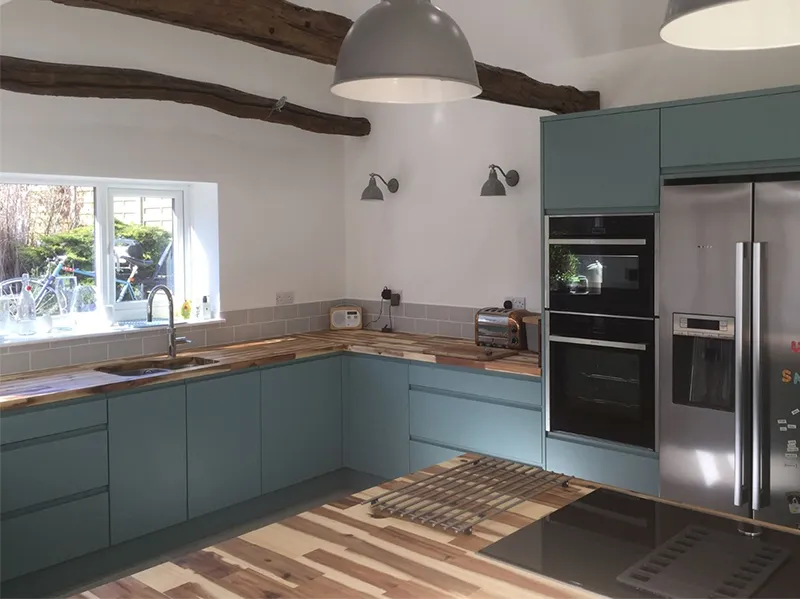 Modern kitchen with teal cabinets, wooden countertops, stainless steel appliances, exposed wooden beams, and large windows letting in natural light.