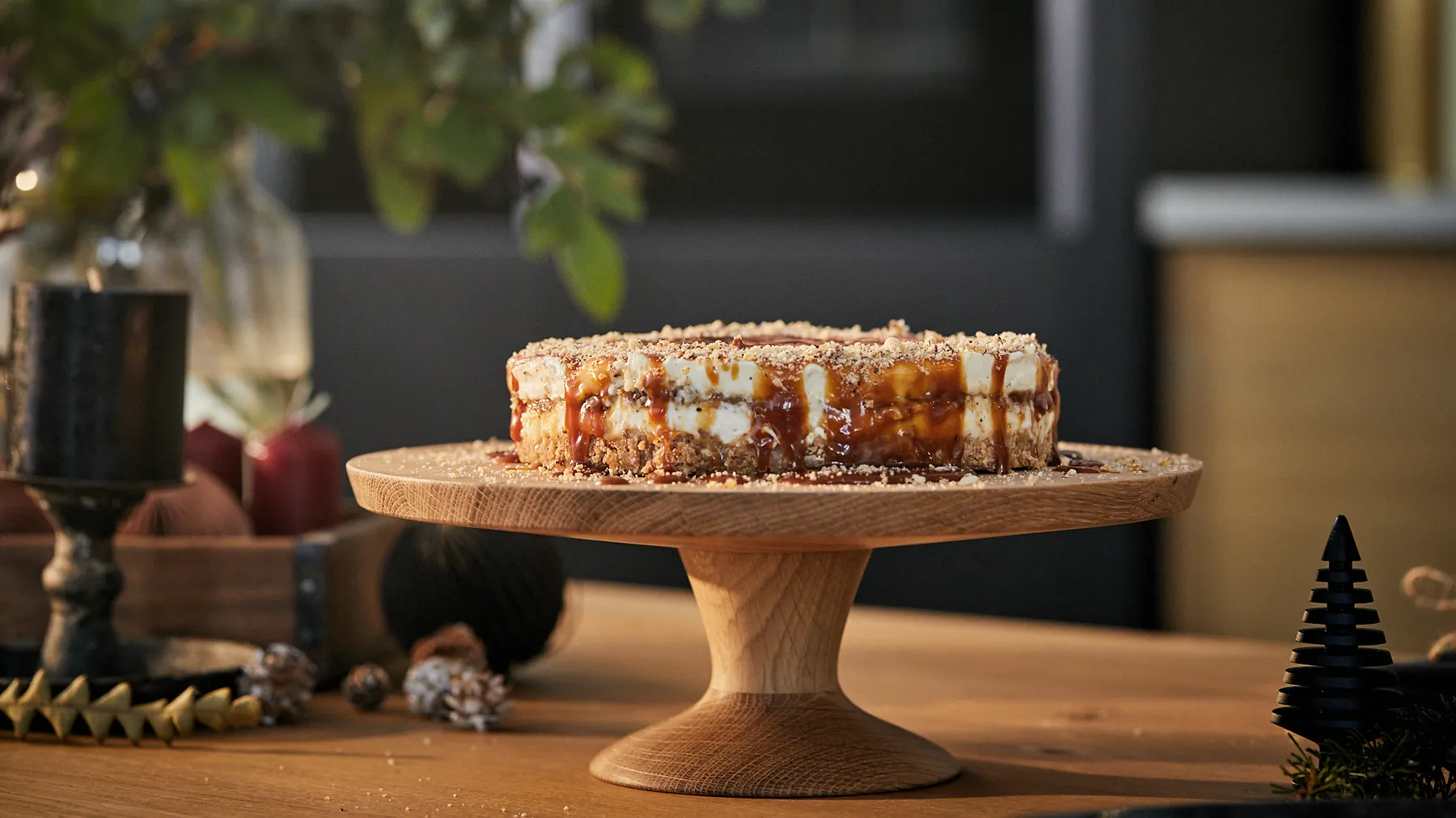 Stand di torta in legno rustico con una torta a strati con glassa e decorazioni, circondato da decorazioni per le vacanze.