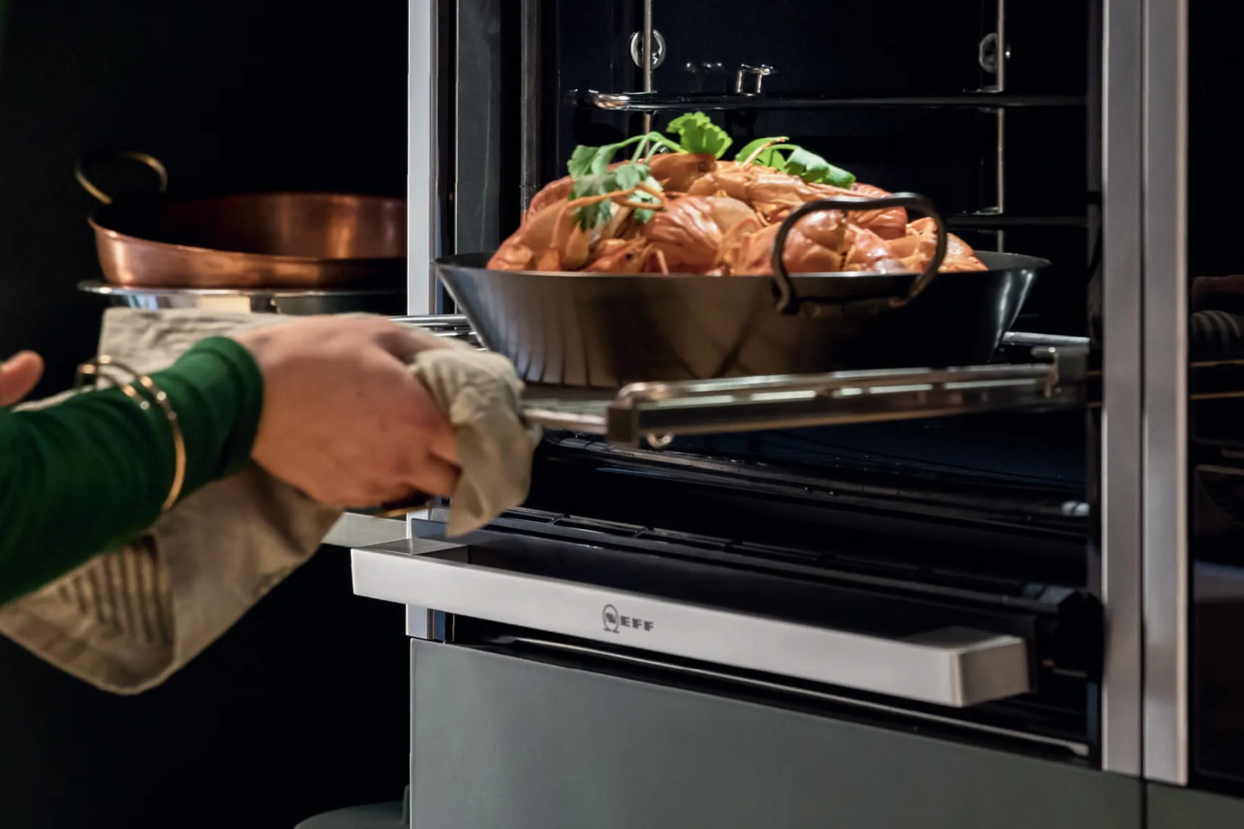 A person wearing oven mitts removes a roasting pan filled with cooked food from a built-in oven.