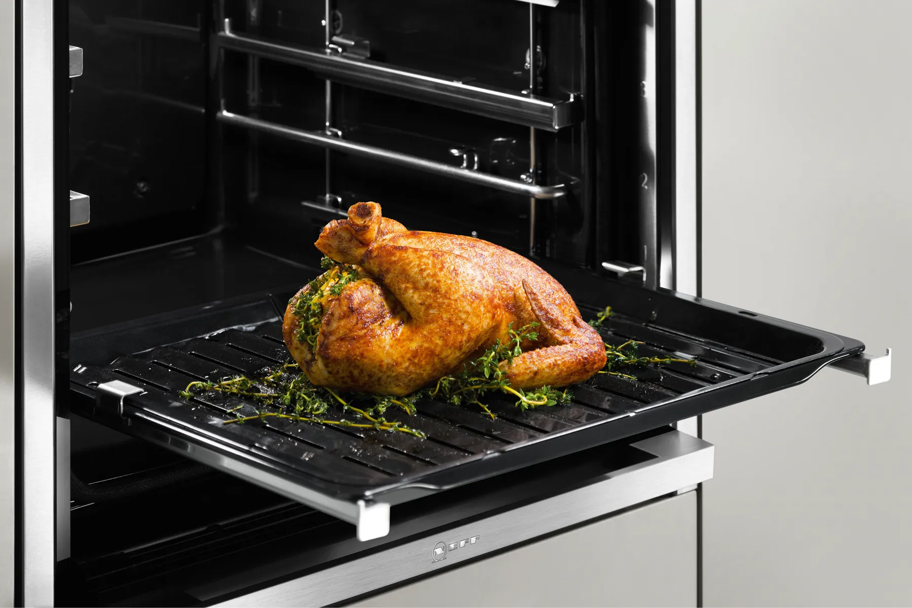 Whole roast chicken with herbs on a roasting rack and tray inside an open stainless-steel oven, oven light illuminating the dish.