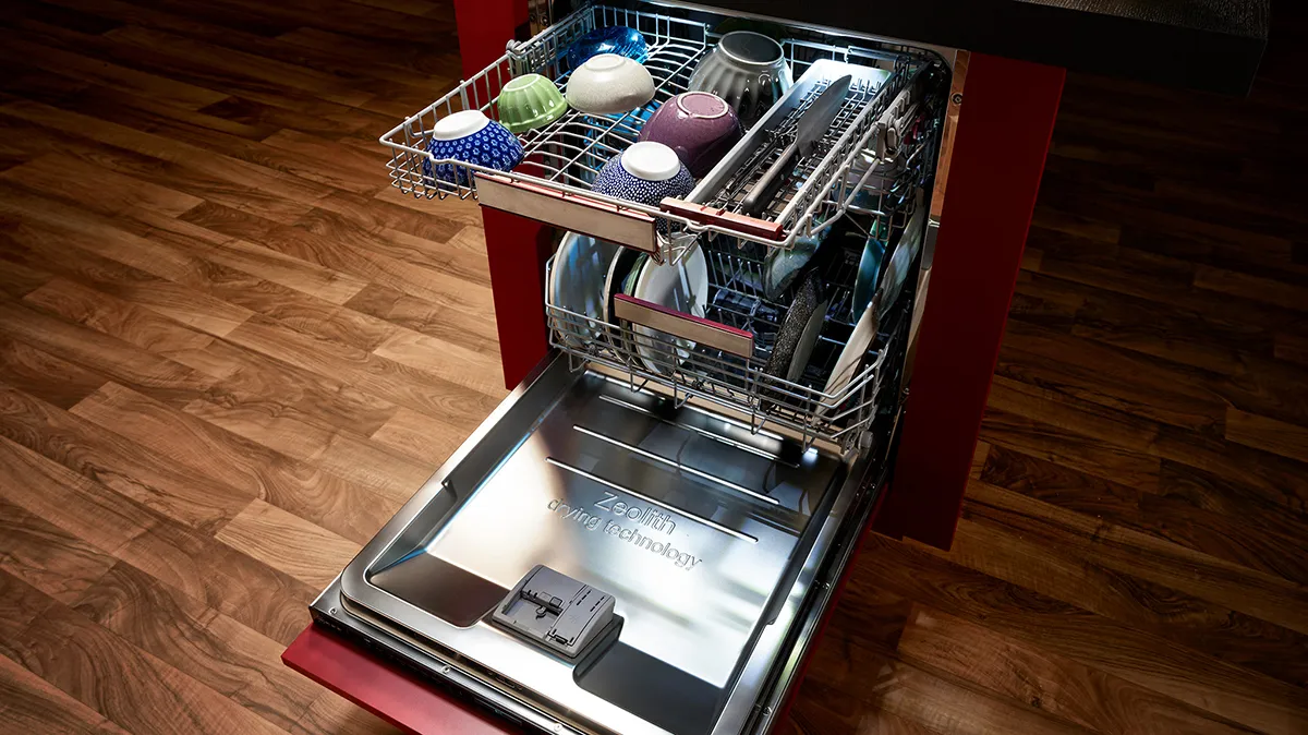 Open dishwasher with dishes, utensils, and cookware inside on wooden floor.