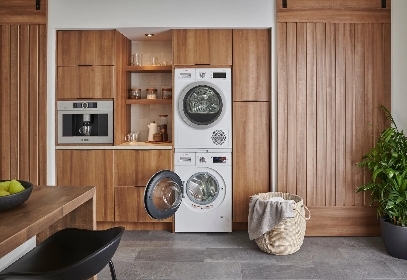 Bosch washer and dryer stacked in a wooden kitchen with an oven, open door, and laundry basket.