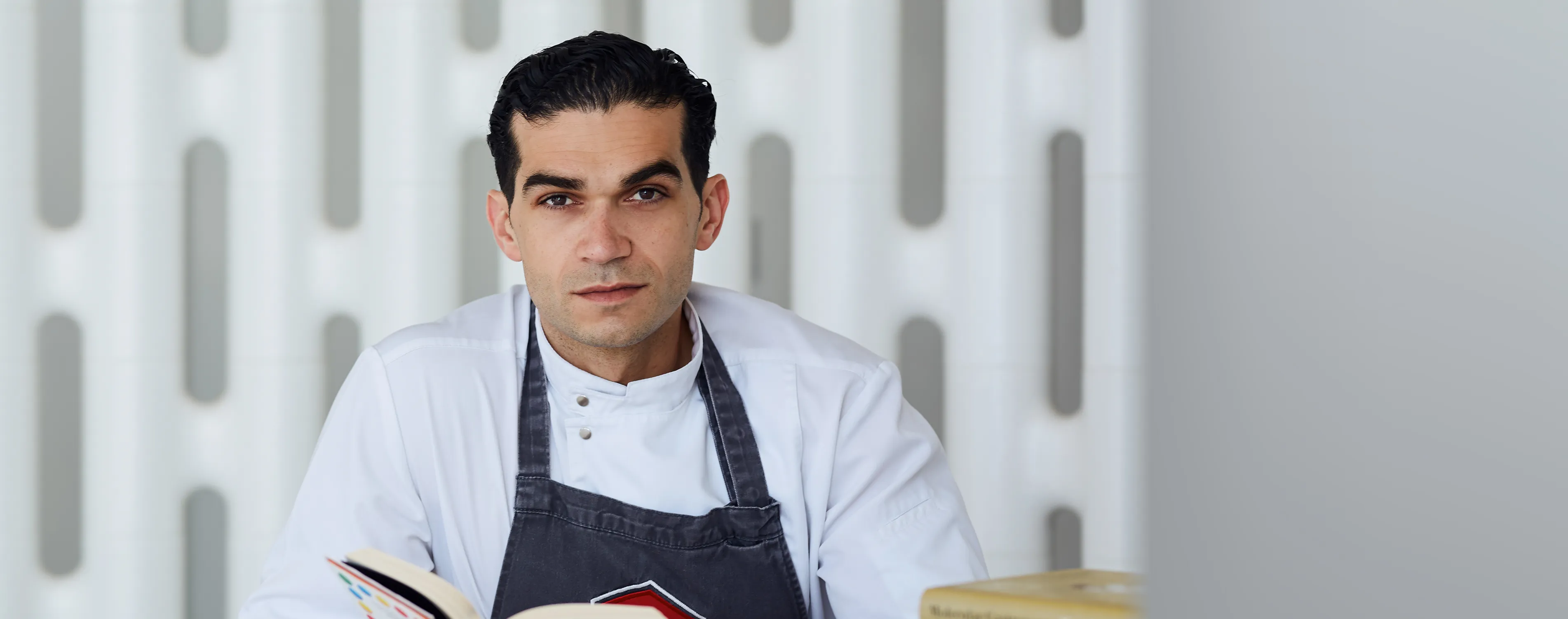 The image shows a young male chef wearing a white uniform and apron, holding a cookbook, standing in a kitchen setting.
