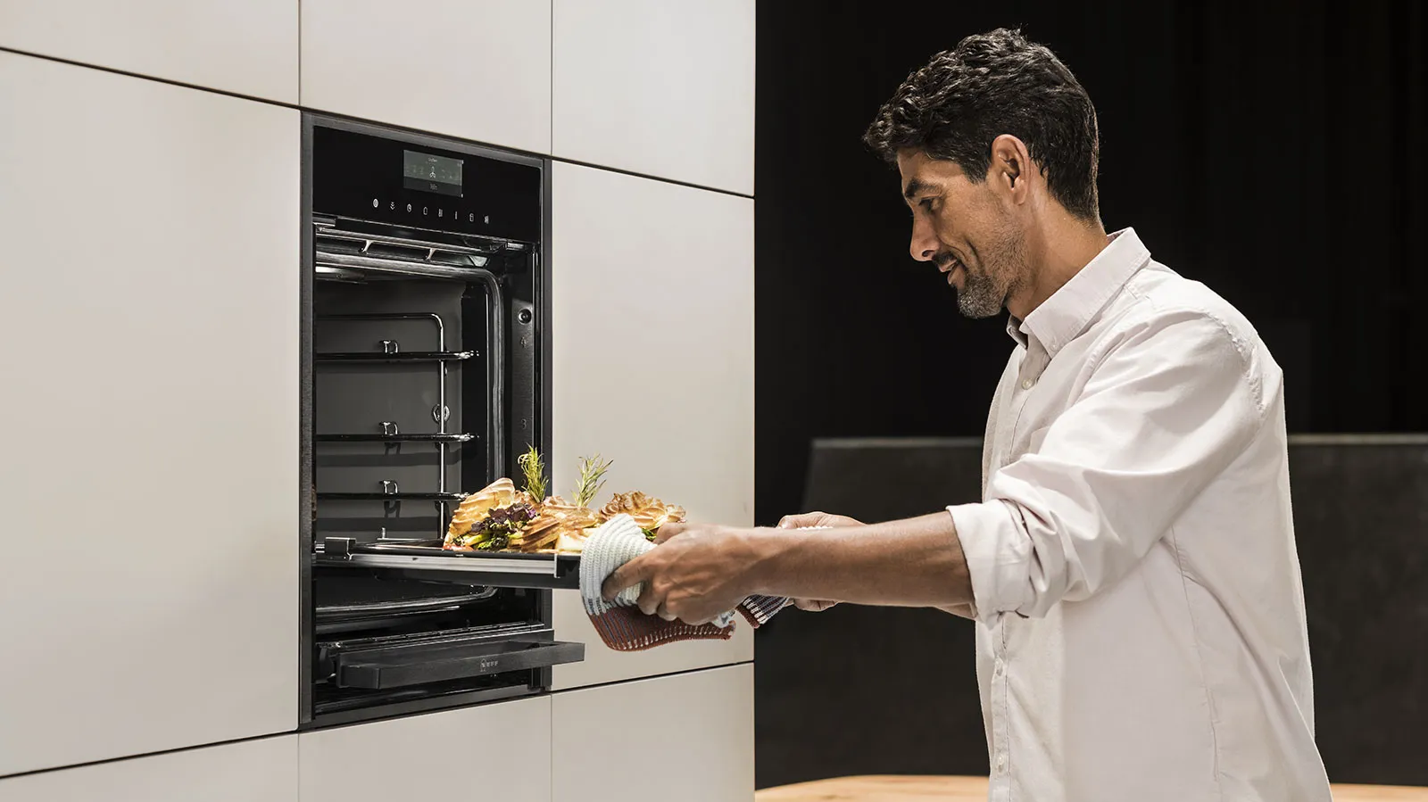 Uno chef estrae con un sorriso una teglia fumante dal forno NEFF, la cui porta a scomparsa Slide & Hide® è completamente rientrata per un accesso comodo e diretto.