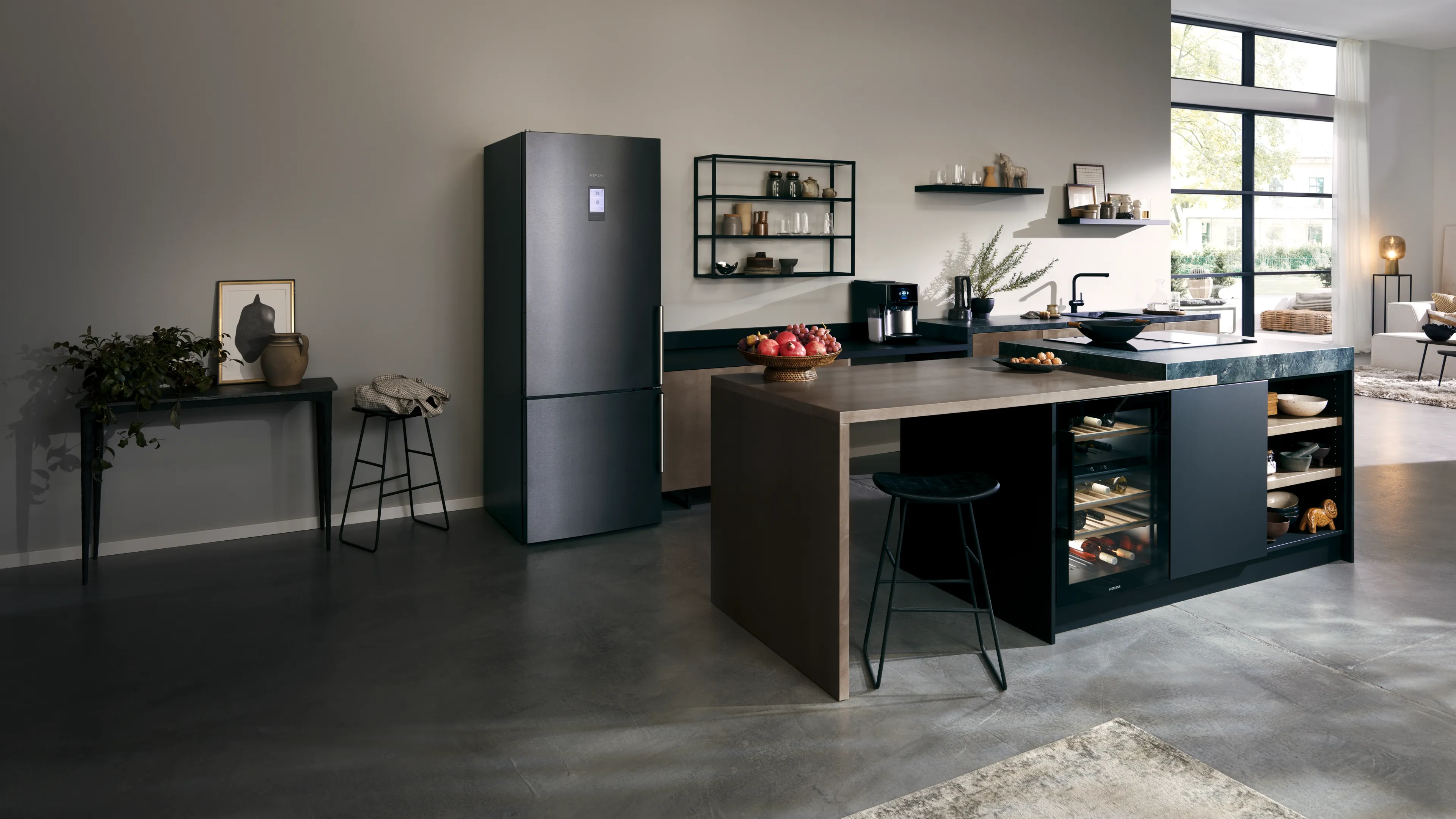 A wide shot of a modern, stylish kitchen featuring a tall, black Siemens fridge-freezer and a large central island. The island has a dark countertop, a built-in wine cooler, and an attached breakfast bar with stools. The open-plan room has concrete floors and opens up to a bright living area in the background.
