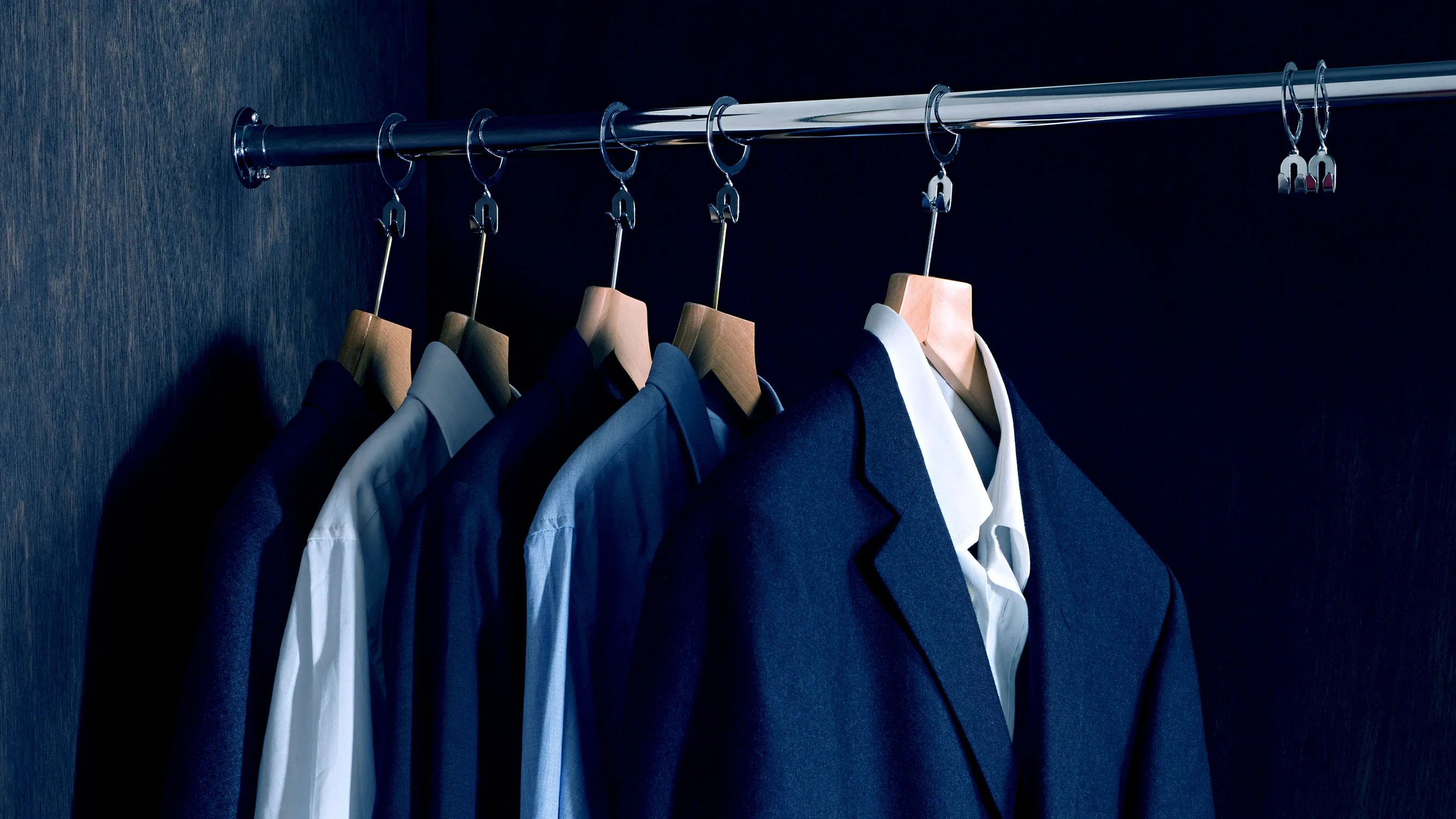 Row of formal suits hanging on a clothing rack against a dark background.