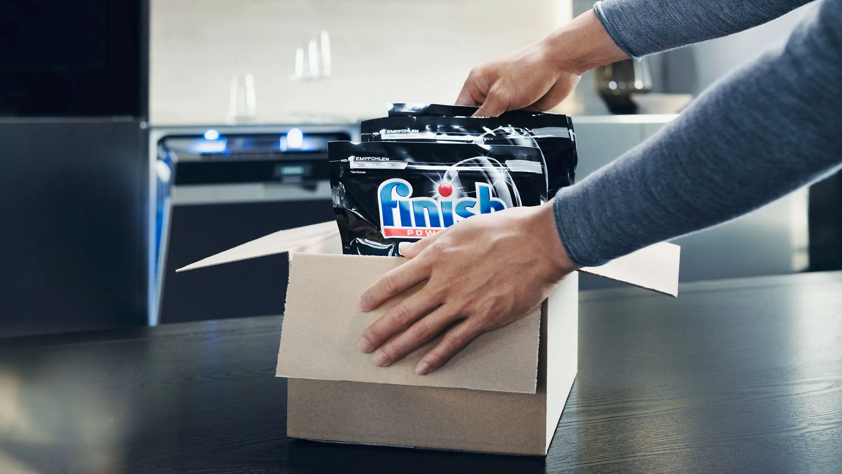Hands packing a printer cartridge into a cardboard box on a desk.