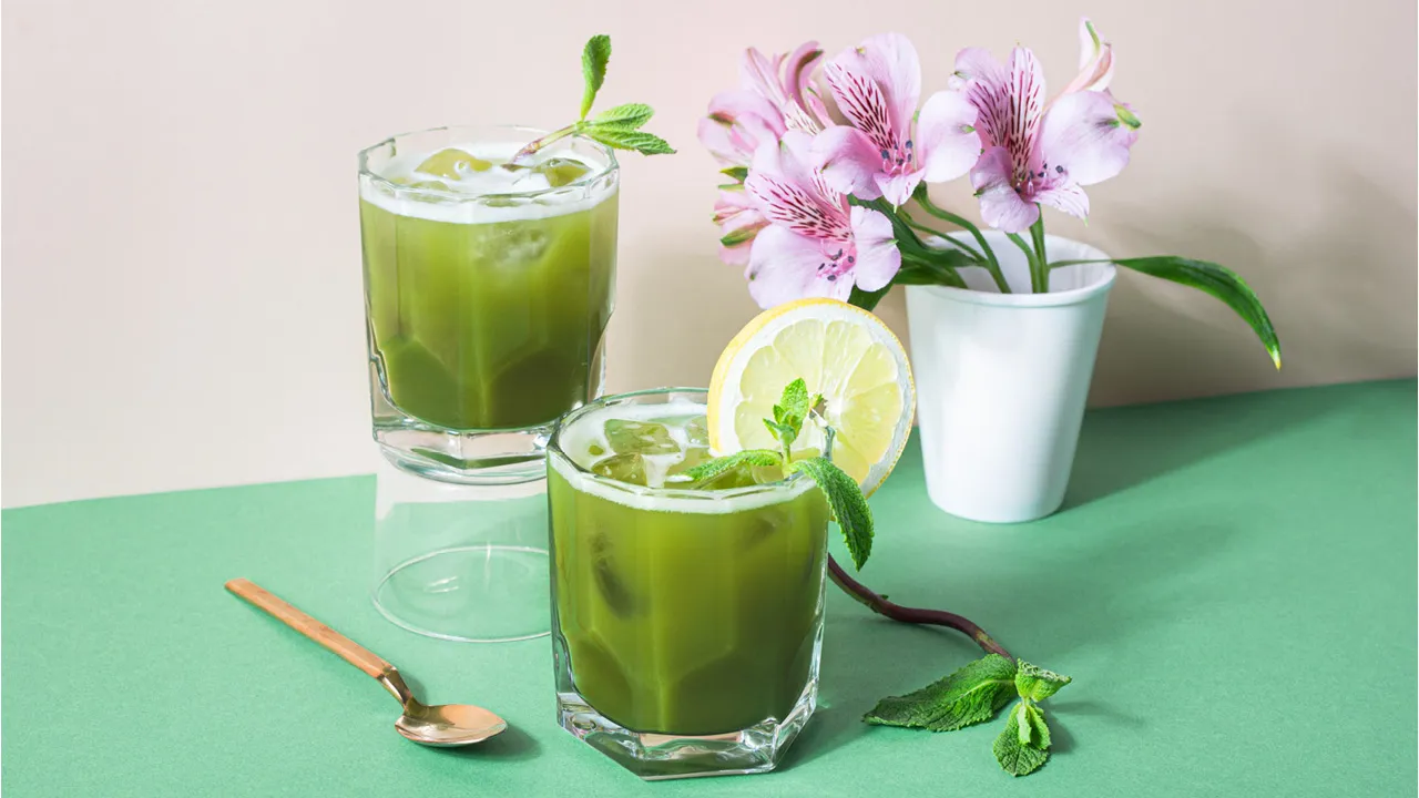 Two glasses of iced matcha with mint and lemon, next to pink flowers in a white pot.